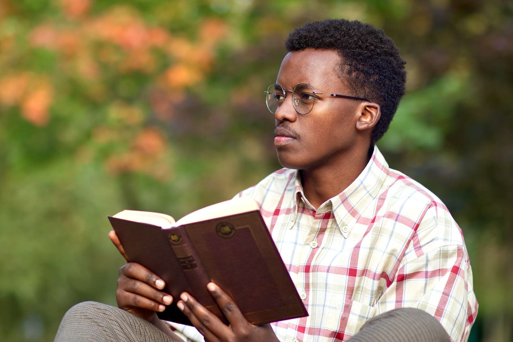 A young man in glasses reading a book outdoors, surrounded by autumn leaves