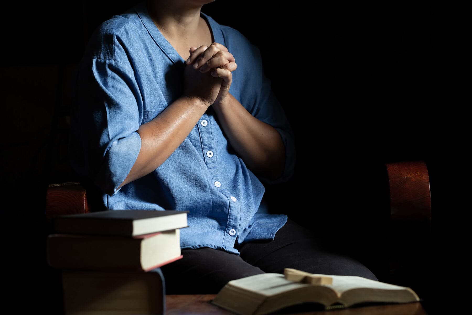 A woman praying with her hands clasped in a dark room, an open Bible on the table in front of her
