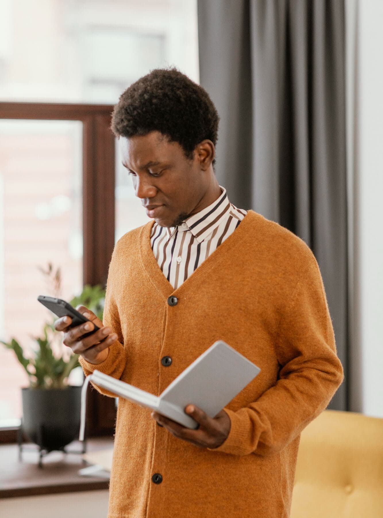 A man in an orange cardigan standing by a window, holding a phone in one hand and a book in the other