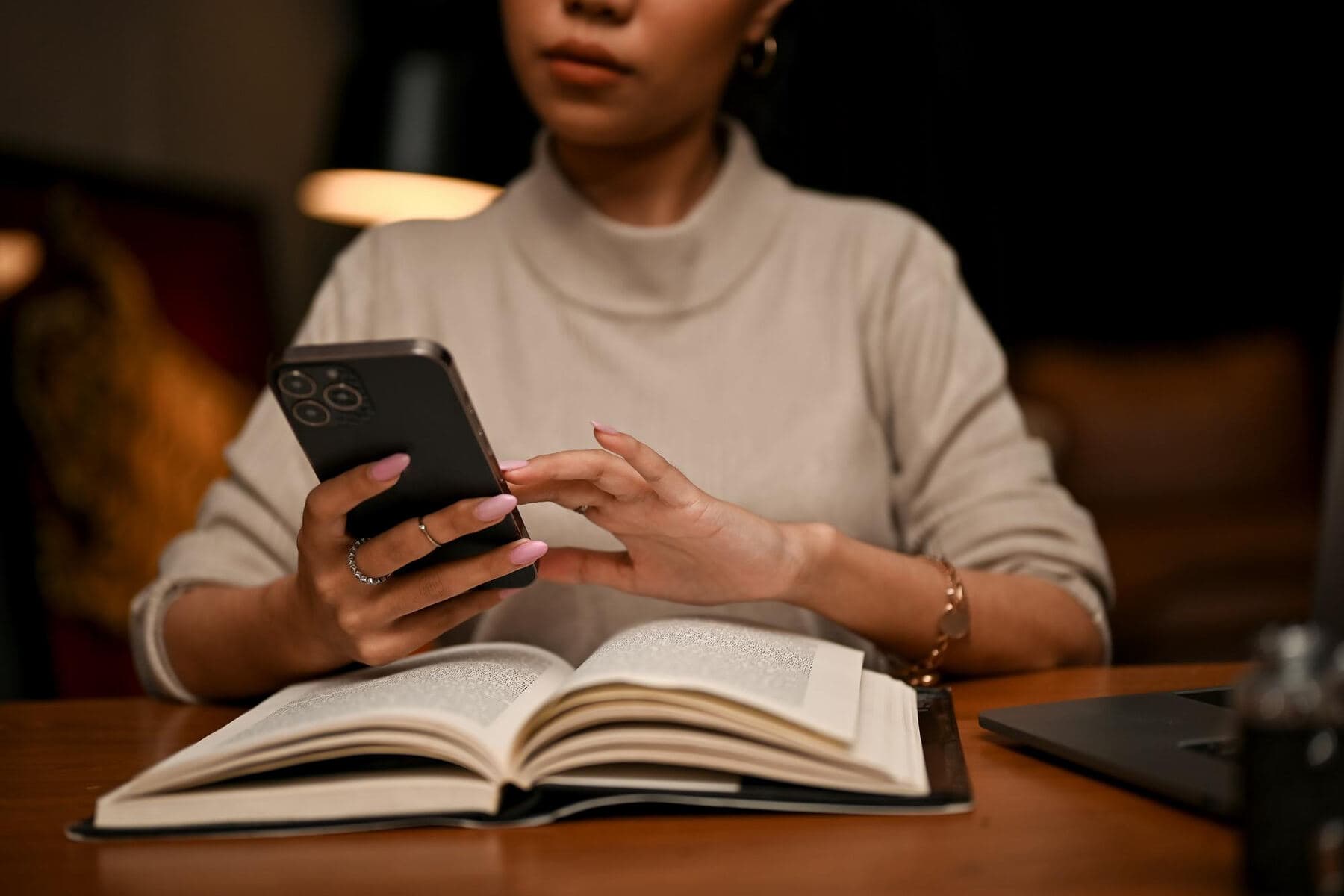 A woman reading on her phone at night beside a lamp, an open book in front of her