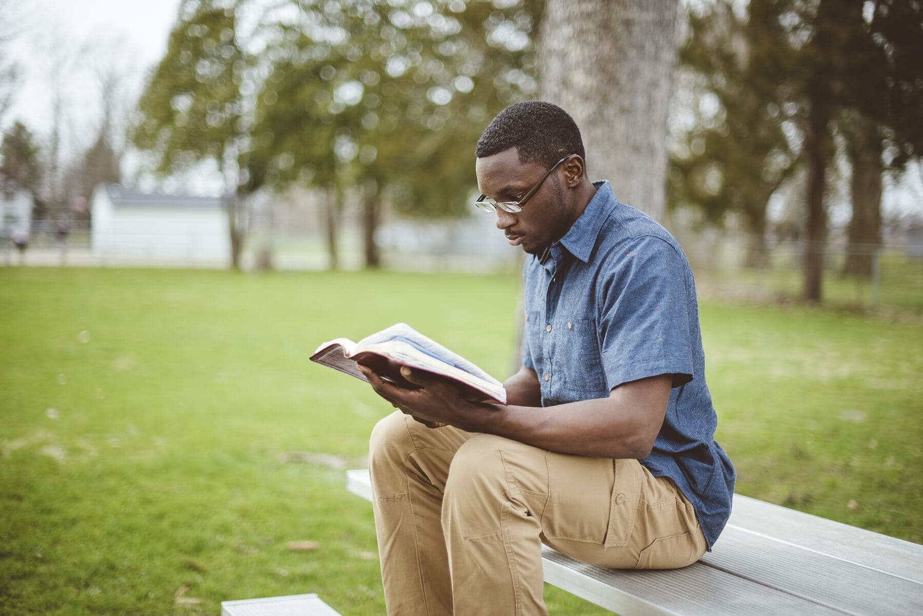 A man sitting on a bench in a park, reading an open Bible