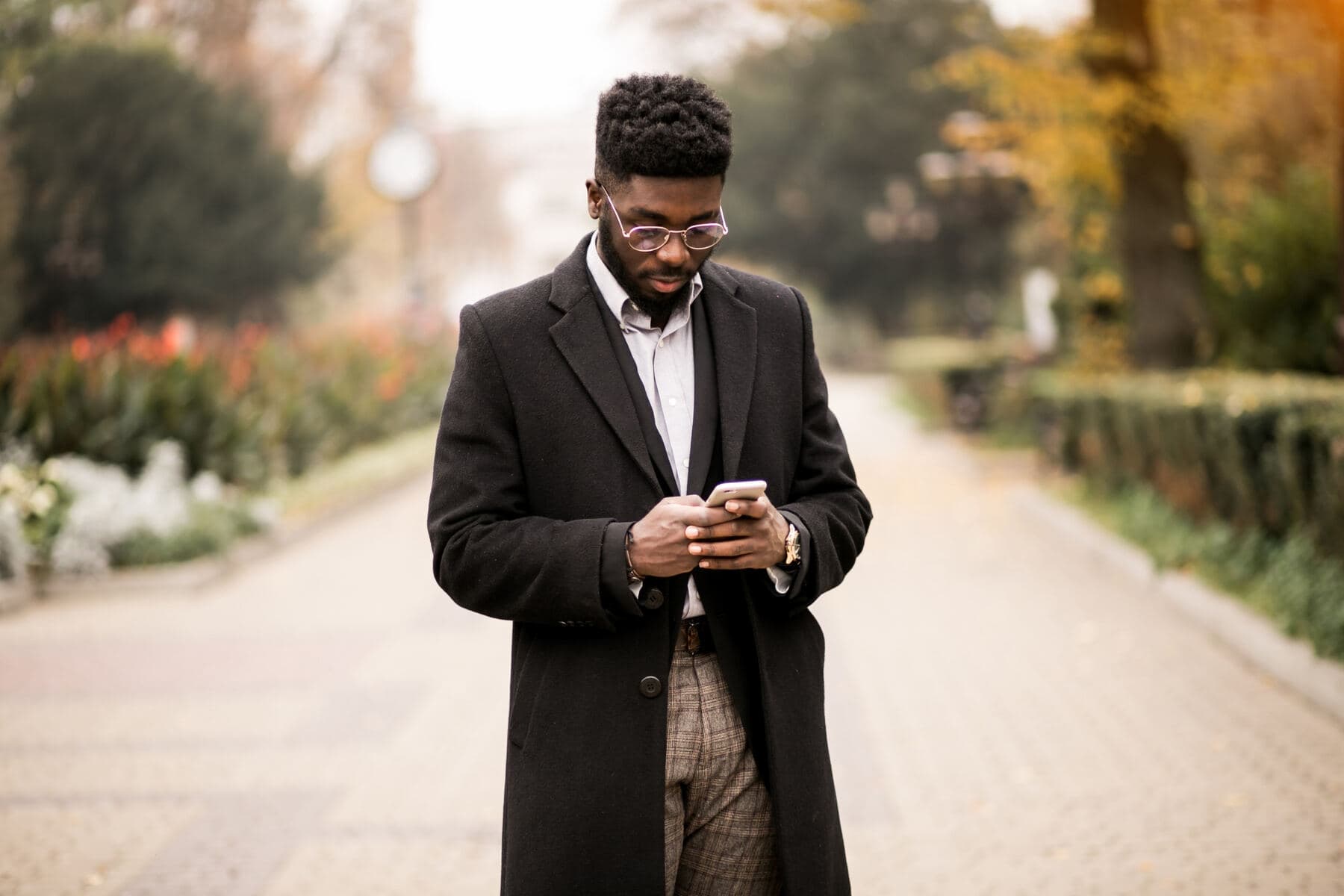 A man walking along a tree lined path, reading something on his phone